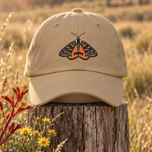 May include: A tan baseball cap featuring an embroidered moth graphic on the front panel. The moth has dark brown wings with intricate white patterns and a vibrant orange section near the body. The cap has a curved brim and a structured crown.