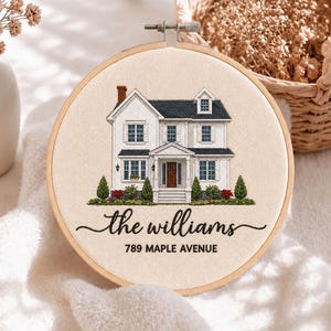 May include: A circular wooden embroidery hoop containing a fabric panel with a stitched illustration of a two-story white house with a dark roof. Below the house, the text reads "the williams" in script and "789 MAPLE AVENUE" in block letters.