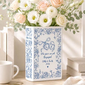 Op de afbeelding: Een rechthoekige witte keramische vaas met een blauw bloemenpatroon en gepersonaliseerde tekst: Colly & Emily, The year we got Engaged, Colly & Emily 2026, inclusief een hartsymbool en twee hartvormige linten met de initialen C en E.