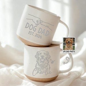 May include: Two speckled off-white ceramic mugs stacked vertically. The top mug features a line drawing of a fist bumping a dog paw with the text DOG DAD EST. 2026. The bottom mug displays a line drawing of a dog with the name Charlie below it.