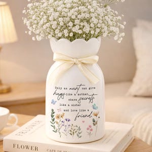May include: A white ceramic vase with a scalloped rim, tied with a cream-colored ribbon, and filled with white flowers. The vase has a floral design and the text "Only an aunt can give hugs like a mother, share secrets like a sister and love like a friend."