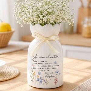 May include: A white ceramic vase with a scalloped rim, tied with a cream ribbon, holds white flowers. The vase features floral illustrations and the text "A new chapter". The vase is decorated with floral illustrations and the text "A new chapter".