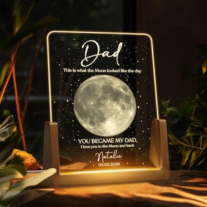K&ouml;nnte beinhalten: Personalisiertes Nachtlicht mit Holzsockel. Die transparente Acrylplatte zeigt das Wort "Dad" und ein Vollmondbild. Der Text enth&auml;lt "This is what the Moon looked like the day" und eine personalisierte Nachricht. Das Datum "05.02.2026" ist ebenfalls sichtbar.