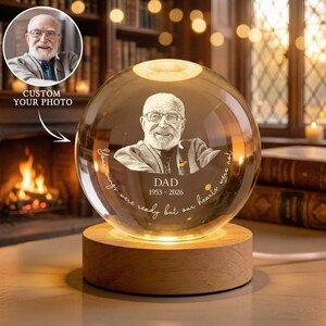May include: A personalized crystal sphere with a photo of a man, the word "DAD", and dates 1953-2026. The sphere sits on a wooden base that emits a warm glow. Text reads "Your wings were ready but our hearts were not."