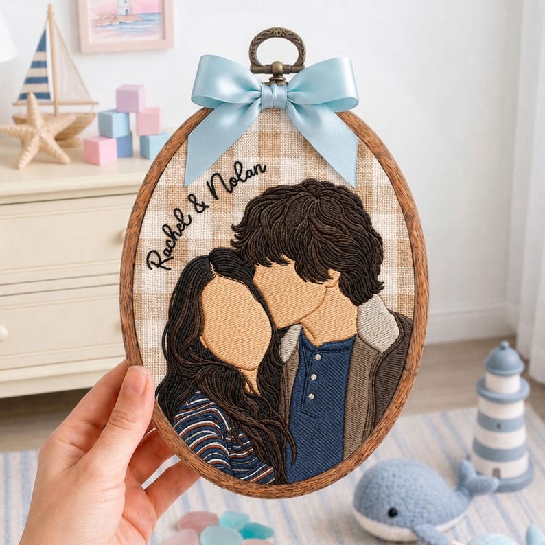 May include: An oval embroidery hoop featuring a stitched portrait of two people with brown hair. The fabric background is a beige and white gingham pattern. The text "Rachel & Nolan" is embroidered in black. A light blue satin bow is attached at the top.
