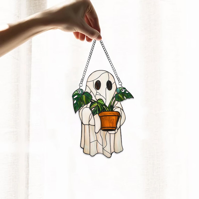 May include: A stained glass ghost holding a potted plant with green leaves. The ghost is white with a light brown face and two black eyes. The plant is in a brown pot. The ghost is hanging from a silver chain.