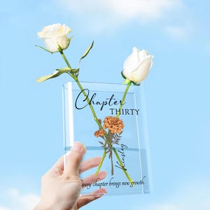 May include: A clear rectangular vase with the words "Chapter Thirty Kinsley" and the phrase "every chapter brings new growth." Two white roses and orange flowers are inside the vase.