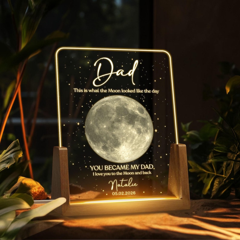K&ouml;nnte beinhalten: Personalisiertes Nachtlicht mit Holzsockel. Die transparente Acrylplatte zeigt das Wort "Dad" und ein Vollmondbild. Der Text enth&auml;lt "This is what the Moon looked like the day" und eine personalisierte Nachricht. Das Datum "05.02.2026" ist ebenfalls sichtbar.