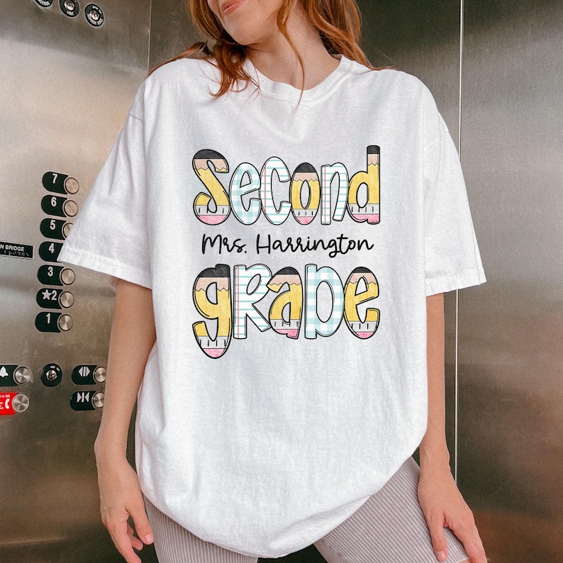 以下が含まれることがあります： 「Second Mrs. Harrington Grade」の文字がカラフルな鉛筆をテーマにしたデザインでプリントされた白いTシャツ。テキストは遊び心のあるフォントで、教師や生徒に最適です。