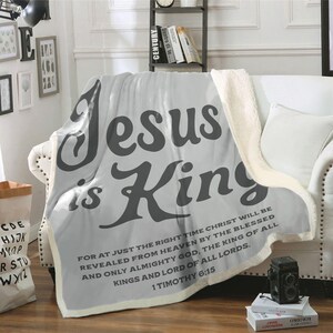 Może przedstawiać: Szara narzuta z napisem "Jesus is King" dużymi, ciemnoszarymi literami, narzucona na białą sofę. Narzuta ma kremową podszewkę z sherpy i cytat z Biblii.