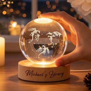 Op de afbeelding: Een heldere glazen bol met drie draken silhouetten en een sterrenachtergrond, van binnenuit verlicht. De bol staat op een houten voet met de inscriptie "Michael's Library". De voet en bol stralen een warme gloed uit.