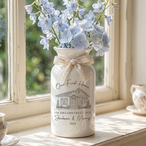 May include: A white ceramic vase featuring a black line drawing of a house, the text "Our First Home", "156 DREAMSTREET AVE", "Jackson & Marry", and "2026". A cream-colored lace ribbon is tied around the neck, holding light blue delphinium flowers.