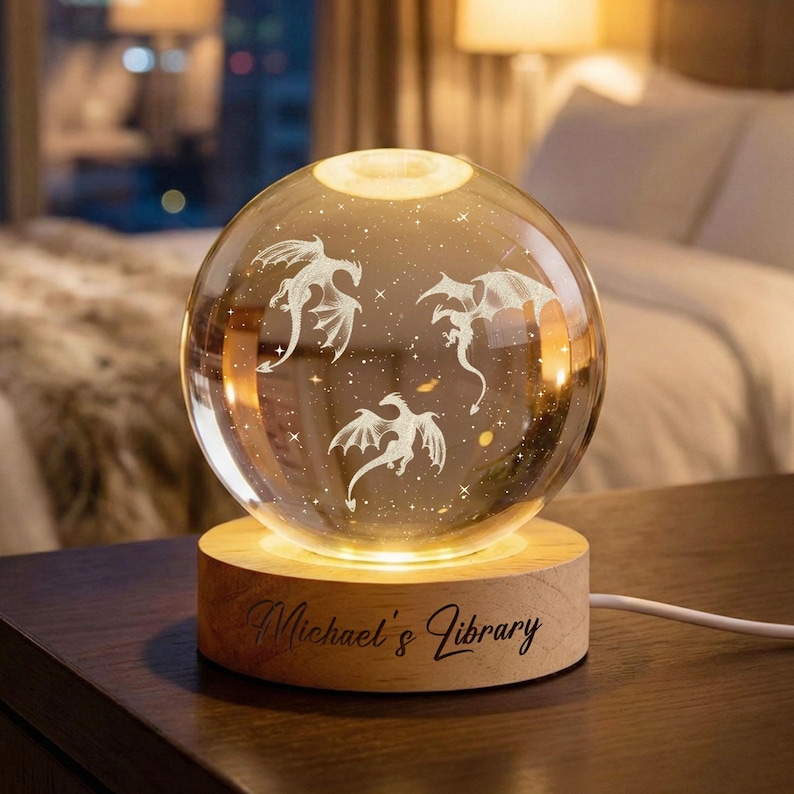 Op de afbeelding: Een decoratieve glazen bol met drie ge&euml;tste drakenontwerpen, verlicht door een warm licht. De bol staat op een houten voet met de inscriptie "Michael's Library". Een snoer loopt van de basis.