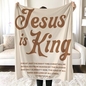 Może przedstawiać: Kremowy koc z napisem "Jesus is King" dużymi, brązowymi, stylizowanymi literami. Poniżej znajduje się werset biblijny z 1 Tymoteusza 6:15. Koc jest trzymany przez osobę.