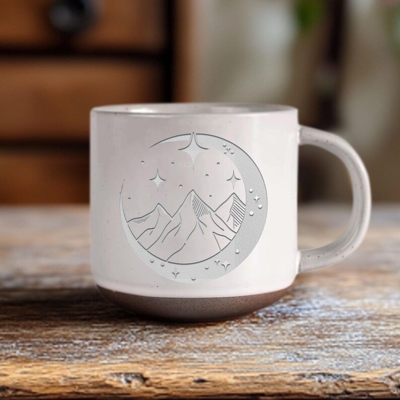 K&ouml;nnte beinhalten: Wei&szlig;e Keramiktasse mit grauer Basis. Die Tasse zeigt eine Schwarzwei&szlig;illustration von Bergen, einem Halbmond und Sternen.