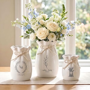 May include: Three white ceramic vases with ruffled rims and tied rope accents. The left vase features a laurel wreath with initials LM and names Larry & Mary. The center vase shows a leaf sprig and Sophia & Henry 09.06.26. The right vase displays a tree.