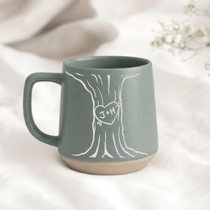 May include: A teal ceramic mug with a tree design and a heart with the initials "J+H". The mug has a speckled finish and a tan base. The handle is square.