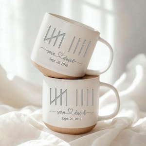 May include: Two white ceramic mugs stacked. Each mug has a light brown base and a white body with a handle. The mugs are decorated with vertical lines, the names "sara & david" with a heart, and the date "Sept. 20, 2016".
