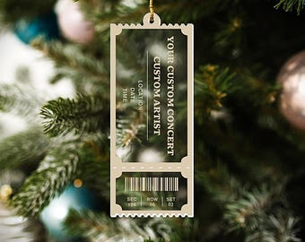 May include: A beige, ticket-shaped Christmas ornament with the text "YOUR CUSTOM CONCERT" and space for customization. The ornament hangs from a gold string and is set against a blurred Christmas tree background.