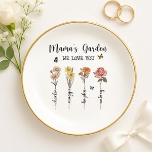 Puede incluir: Plato de cer&aacute;mica blanca con borde dorado. El plato presenta un dise&ntilde;o floral con el texto "Mama's Garden We Love You" y cuatro nombres escritos verticalmente debajo de las flores.