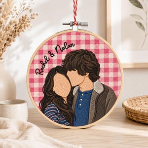 May include: A circular embroidery hoop featuring a stitched portrait of two people against a pink and white gingham background. The text "Rachel & Nolan" is embroidered in black script. The hoop is suspended by a red and white twisted cord.