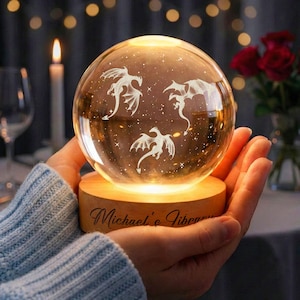 Op de afbeelding: Een heldere glazen bol met drie ge&euml;tste drakenontwerpen, van binnenuit verlicht, rust op een houten voet. De voet is gegraveerd met "Michael's Library". De bol wordt in samengevouwen handen gehouden.