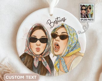 May include: A round, white ornament featuring an illustration of two women wearing sunglasses and headscarves. The word "Besties" is written above the image. A small photo with the text "UPLOAD PHOTO" is in the upper right corner. The text "CUSTOM TEXT" is at the bottom.