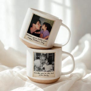 May include: Two white ceramic mugs with a light brown base are stacked. Each mug features a vintage-style photograph. One mug has a photo of a man and child with the text "Forever your little girl. Happy Father's Day." The other mug has a photo of a man and child with the text "Grandpa you're the best Love Emily."