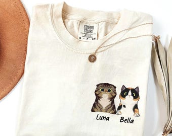 May include: Cream-colored t-shirt featuring two cartoon cats, Luna and Bella, with their names below. The shirt has a Comfort Colors tag and a gold necklace. A brown hat and dried plant are in the background.