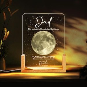 K&ouml;nnte beinhalten: Ein personalisiertes Nachtlicht mit Holzsockel und klarer Acrylplatte. Die Platte zeigt das Wort "Dad", eine Vollmondgrafik und den Text "You became my Dad. I love you to the Moon and back." mit einem Namen und Datum.