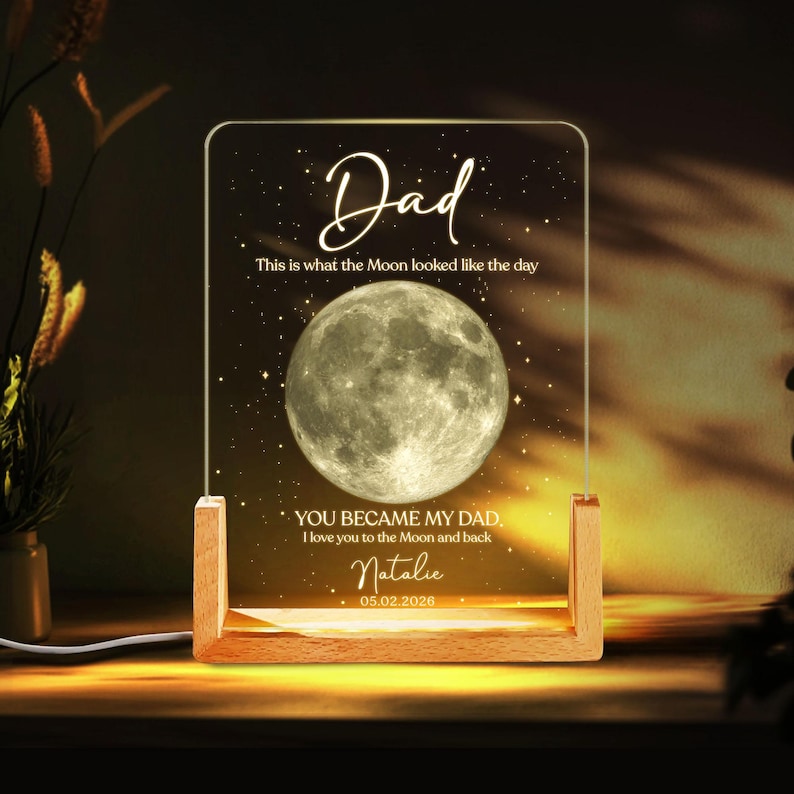 K&ouml;nnte beinhalten: Ein personalisiertes Nachtlicht mit Holzsockel und klarer Acrylplatte. Die Platte zeigt das Wort "Dad", eine Vollmondgrafik und den Text "You became my Dad. I love you to the Moon and back." mit einem Namen und Datum.