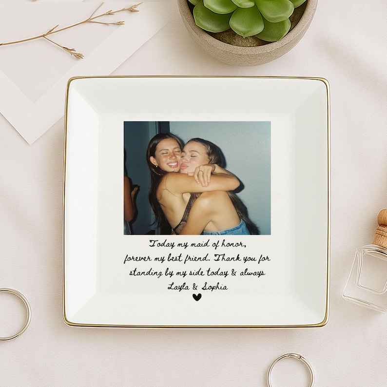 May include: A square white ceramic trinket dish with a gold rim. The dish features a photograph of two women embracing. Below the photo is handwritten text that reads "Today my maid of honor, forever my best friend..."