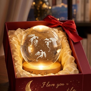 Op de afbeelding: Een heldere glazen bol met drie ge&euml;tste drakenontwerpen, van binnenuit verlicht, staat op een houten voet in een rode geschenkdoos. De doos is gevoerd met gouden stof en vastgebonden met een rode strik. De tekst "I love you" is zichtbaar.