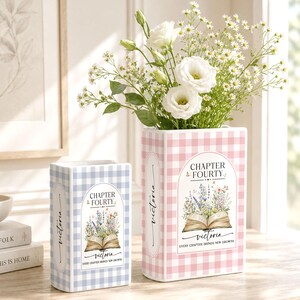 May include: Two rectangular ceramic vases shaped like books with a gingham pattern. The smaller vase is light blue, and the larger pink vase holds white flowers. Both feature the text "CHAPTER FOURTY", "victoria", and "EVERY CHAPTER BRINGS NEW GROWTH" with an open book and floral illustration.