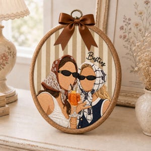 May include: An oval embroidery hoop art piece featuring two figures wearing headscarves and black sunglasses, holding an orange cocktail. The background is beige and white striped fabric with the word Besties embroidered in black, topped with a brown satin bow.