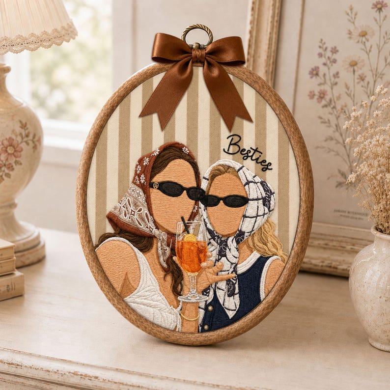May include: An oval embroidery hoop art piece featuring two figures wearing headscarves and black sunglasses, holding an orange cocktail. The background is beige and white striped fabric with the word Besties embroidered in black, topped with a brown satin bow.