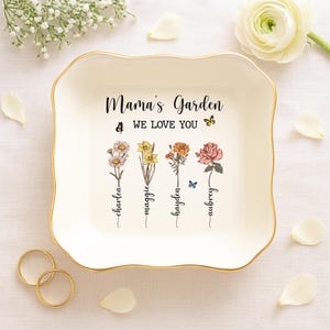 Puede incluir: Plato de cer&aacute;mica blanca para joyas con borde dorado. El plato tiene un dise&ntilde;o floral con el texto "Mama's Garden We Love You" y los nombres "Charlea, Maggie, Hayden, Aubrey" escritos debajo de las flores.