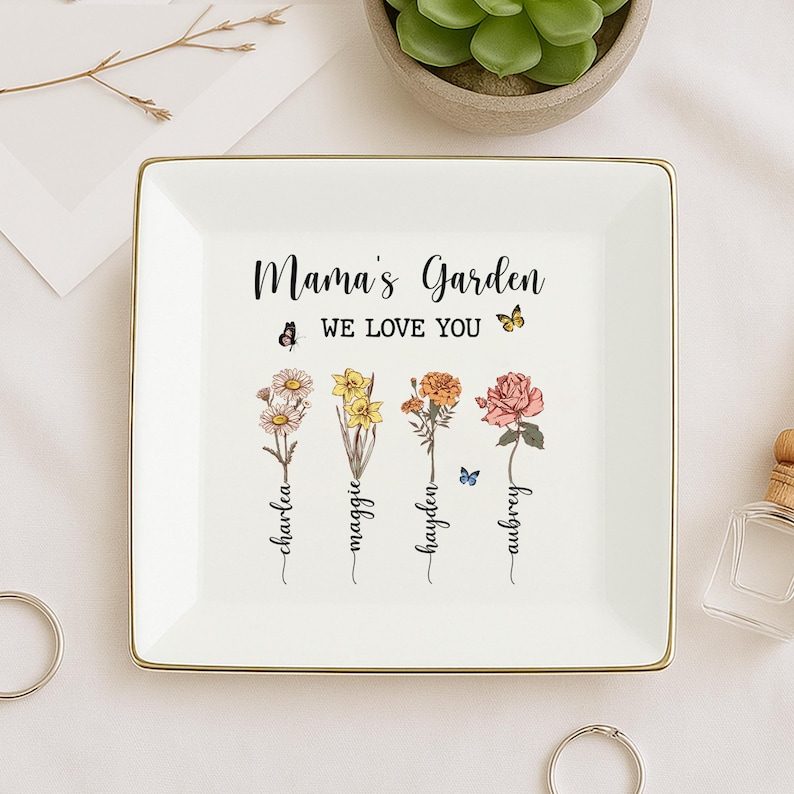 Puede incluir: Bandeja de cer&aacute;mica blanca con borde dorado. La bandeja presenta un dise&ntilde;o floral con el texto "Mama's Garden We Love You" y los nombres "Charlea, Maggie, Hayden, Aubrey" escritos debajo de las flores.