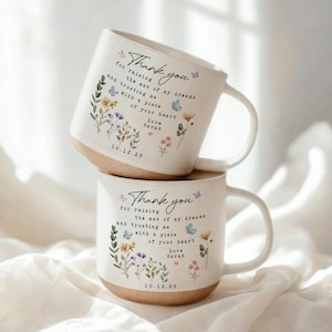 May include: Two white ceramic mugs with a floral design and the text "Thank you" and a personalized message. The mugs have a beige base and a curved handle. The floral design includes butterflies and wildflowers.