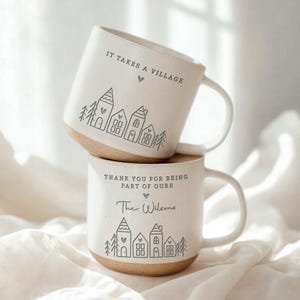 May include: Two off-white ceramic mugs with speckled textures and a handle. The top mug features the text IT TAKES A VILLAGE and a line drawing of houses and trees. The bottom mug features the text THANK YOU FOR BEING PART OF OURS and The Wilsons.