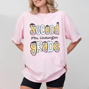 以下が含まれることがあります： 明るいピンク色のTシャツ。カラフルで遊び心のあるフォントで「Second Grade」と書かれています。テキストには「Mrs. Harrington」の名前が含まれています。デザインには鉛筆とクレヨンの要素が組み込まれており、新学期のテーマを作成しています。