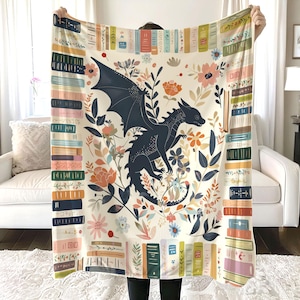 May include: A white throw blanket with a black dragon and floral design. The blanket features a border of colourful stacked books.
