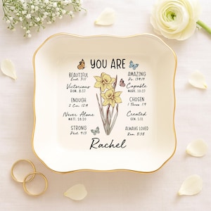 May include: White ceramic trinket dish with a gold rim. The dish features a floral design with yellow daffodils and butterflies. The text "YOU ARE" is printed in black at the top of the dish, followed by a list of positive affirmations with corresponding Bible verses. The name "Rachel" is printed in black at the bottom of the dish.