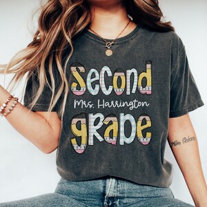 以下が含まれることがあります： 「Second Grade」の文字がカラフルで遊び心のあるフォントでプリントされたダークグレーのTシャツ。「Mrs. Harrington」の名前も含まれています。文字は鉛筆とチェック柄のようにデザインされています。