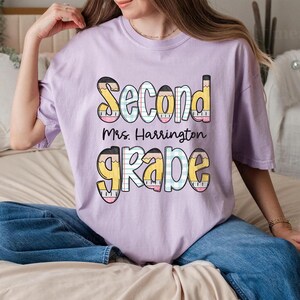 以下が含まれることがあります： カラフルな鉛筆のデザインで「Second Grade Mrs. Harrington」と書かれた、薄紫色のTシャツ。「Second」の文字は黄色とピンク、「Grade」は黄色と青色です。