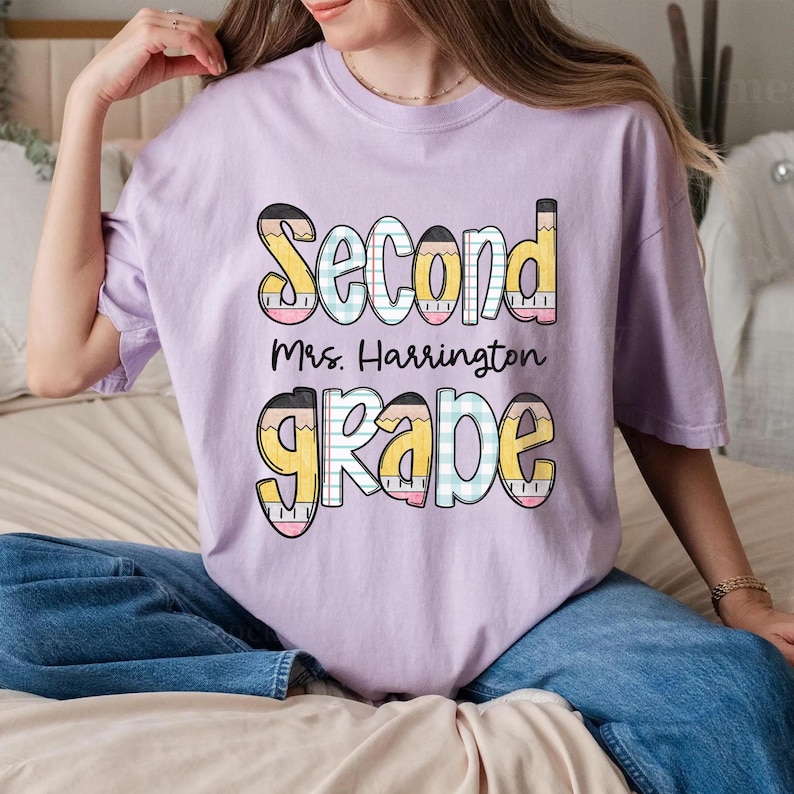 以下が含まれることがあります： カラフルな鉛筆のデザインで「Second Grade Mrs. Harrington」と書かれた、薄紫色のTシャツ。「Second」の文字は黄色とピンク、「Grade」は黄色と青色です。