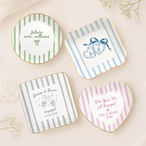 May include: Four ceramic trinket dishes with gold rims and striped patterns. One round green dish says future mrs. williams, one square blue dish features two hearts with letters L and E, one square grey dish says James & Emma engaged 07.15.2025, and one heart-shaped pink dish says The Year We got Engaged Tim & Rachel 2025.