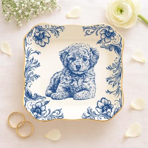May include: A square, off-white decorative plate with a blue illustration of a puppy. The plate has a gold trim and blue floral designs around the edges. The puppy is in the center of the plate.