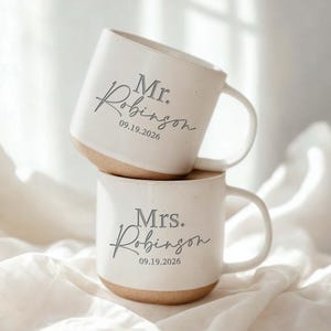 May include: Two off-white ceramic mugs with speckled textures and tan bases. The top mug features the text Mr. Robinson 09.19.2026, and the bottom mug features the text Mrs. Robinson 09.19.2026 in a dark gray script and sans-serif font combination.