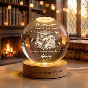 May include: A clear glass globe with a wooden base, engraved with graduation details. The globe features the text "Mastered it!" and "University of Michigan." It also includes the name "Amelia" and the year "2026."